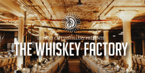 Pictured is a venue in an industrial yet elegant setting at the Whiskey Factory in Detroit. There are string lights, long tables covered with white formal tablecloths, and elegant event decor such as candles and floral centerpieces. The site is prepared to host a corporate event or a wedding reception. Overlayed on top of the image is a logo stating "Detroit City Distillery Presents The Whiskey Factory"